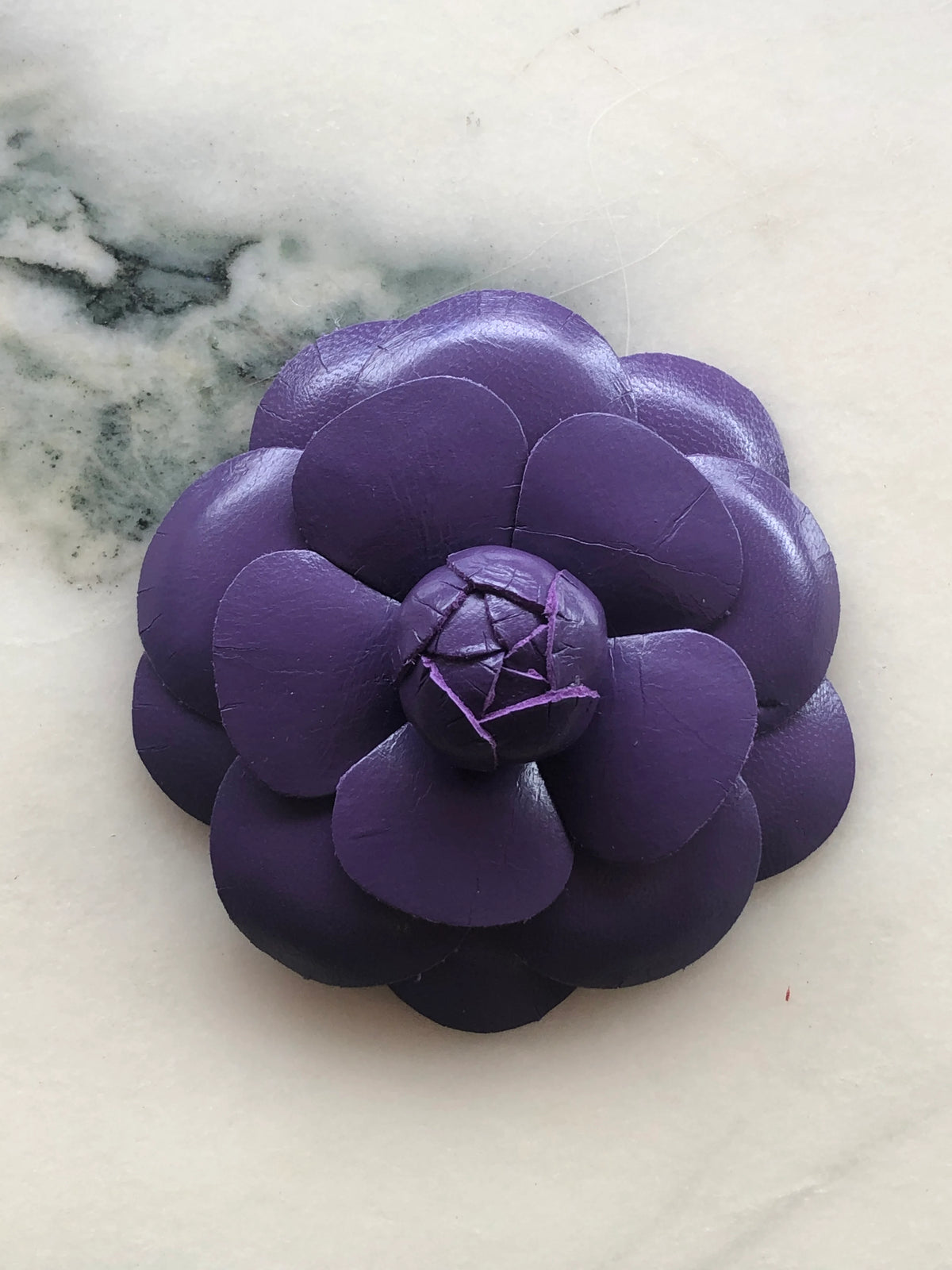 A Broochella Camelia handmade purple leather flower-shaped accessory brooch,  resting on a white marble surface.