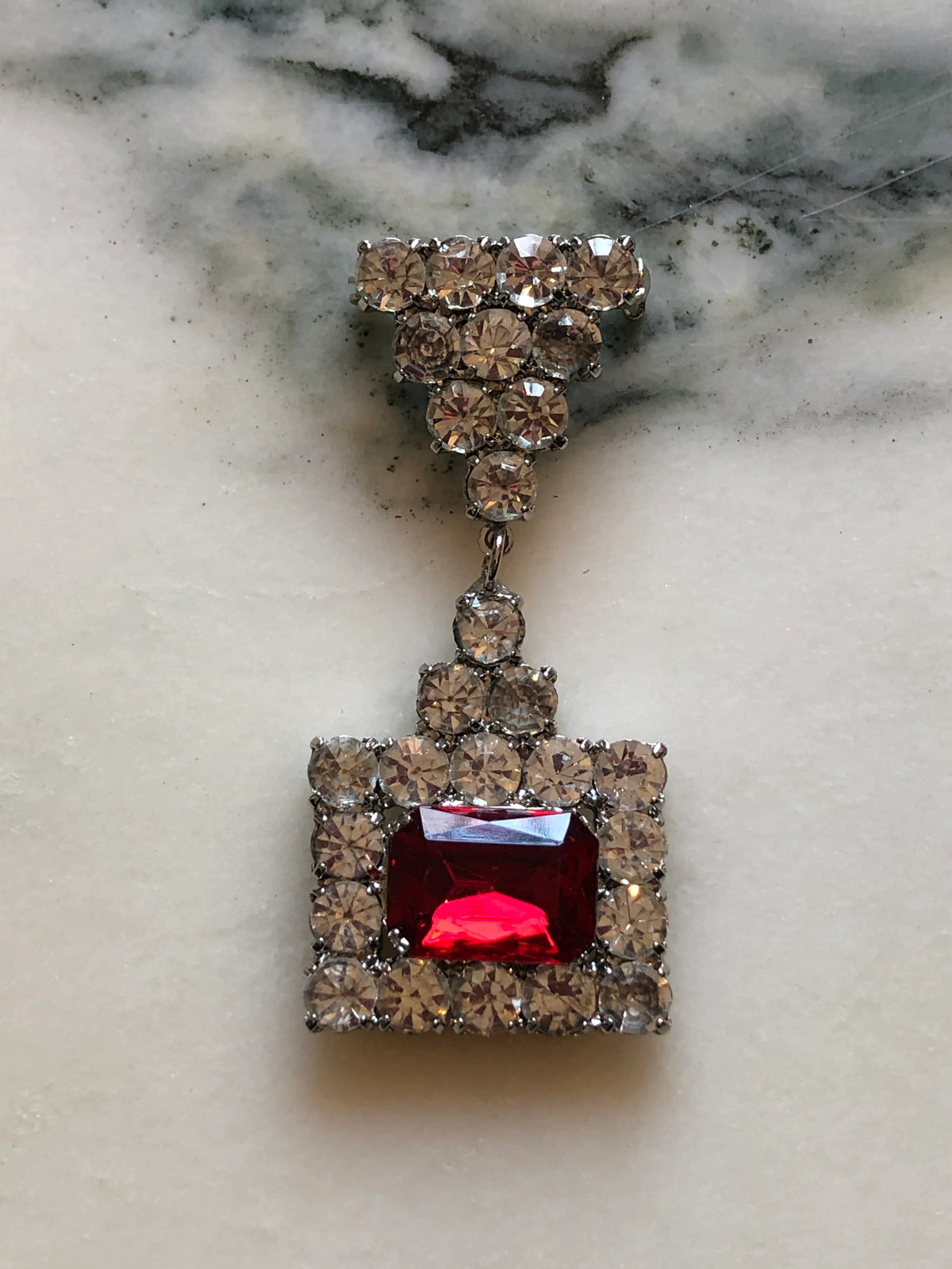 Broochella Raffaella Vintage Red Crystal Drop Brooch featuring a rectangular ruby-red faceted stone framed by clear crystals with an articulated crystal drop, photographed on a cream and grey marble background.