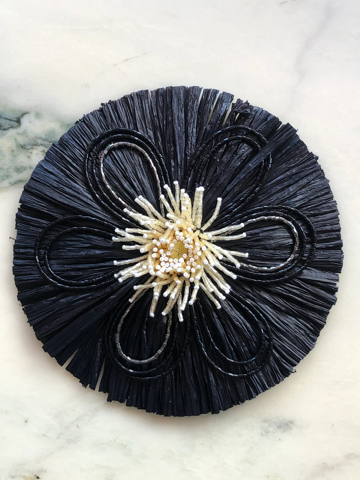 Broochella Raffia Bloom Brooch Pin in black with sculptural raffia fringe, black beaded flower petals and an ivory beaded centre, photographed on a white marble background.