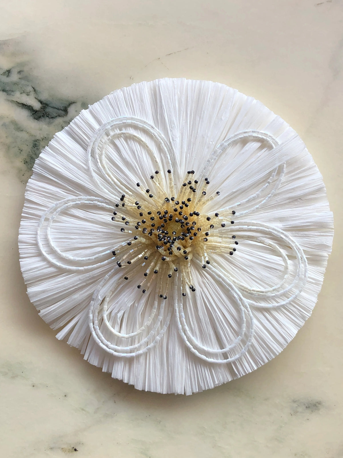 Broochella Men's Raffia Bloom Brooch Pin in white with sculptural raffia fringe, white beaded flower petals and a dimensional ivory beaded centre with black accents, photographed on a white marble background.