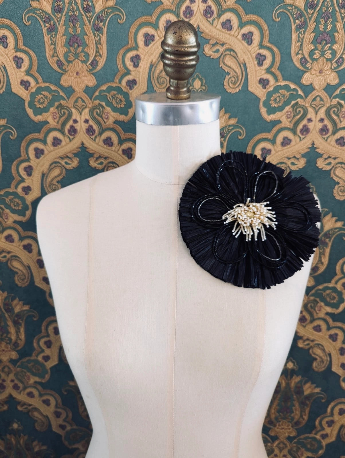 Broochella Raffia Bloom black handmade raffia flower brooch with hand beaded centre. Pinned to a mannequin. The background is green and gold damask wallpaper