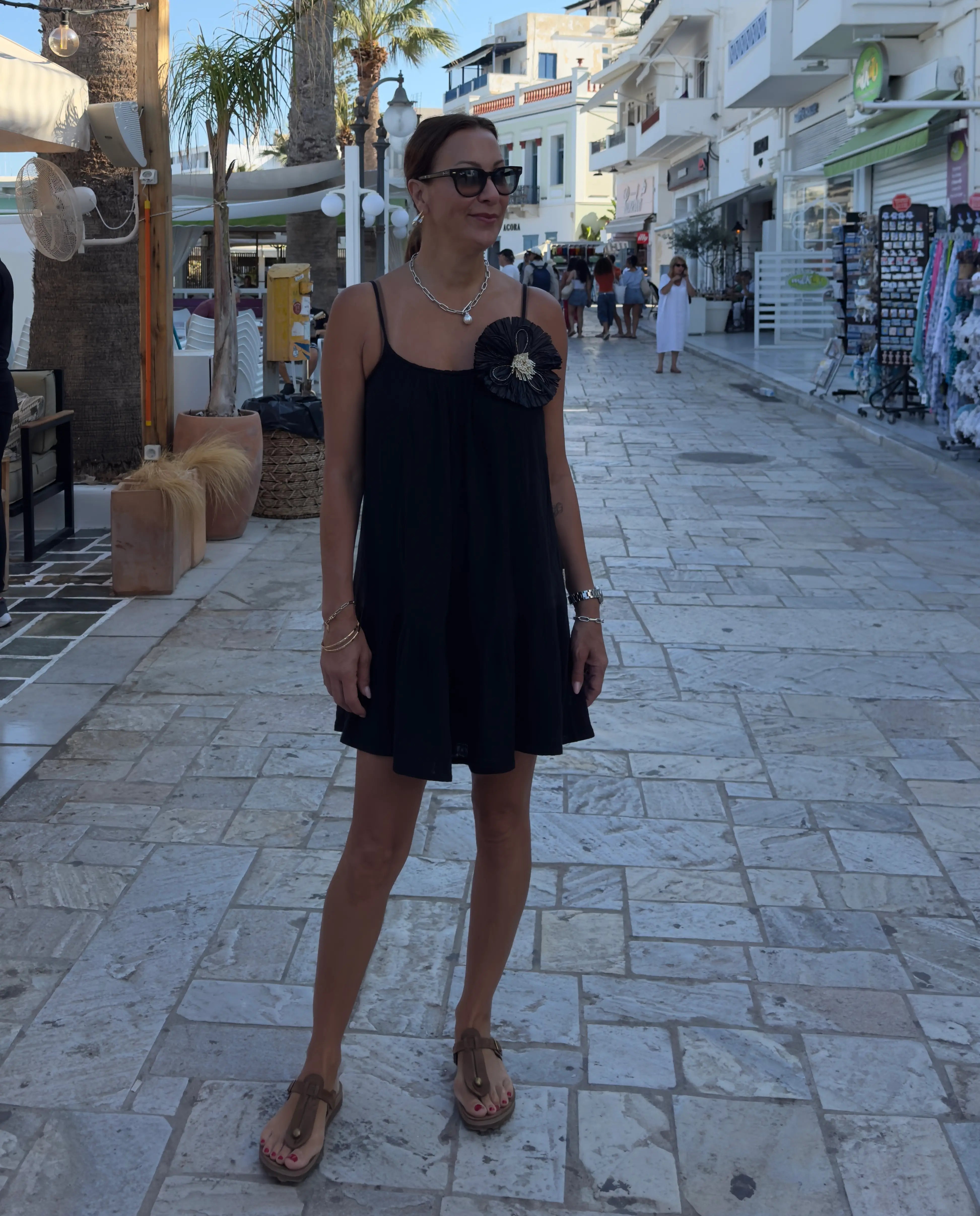 A woman wearing a black sleeveless mini dress with a black floral embellishment on the chest, standing on a stone-paved outdoor street in a Mediterranean-style town. She is wearing the broochella raffia flower brooch in black