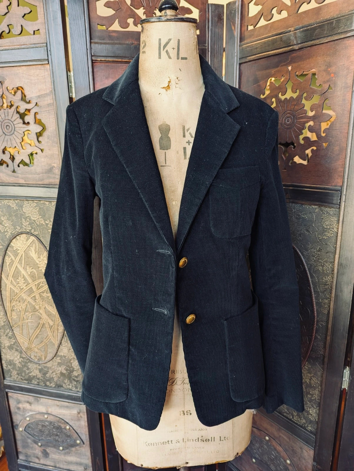 A broochell Rag and Bone  black corduroy blazer jacket displayed on a vintage cream-colorued dress form mannequin, set against a dark wooden carved cabinet background.