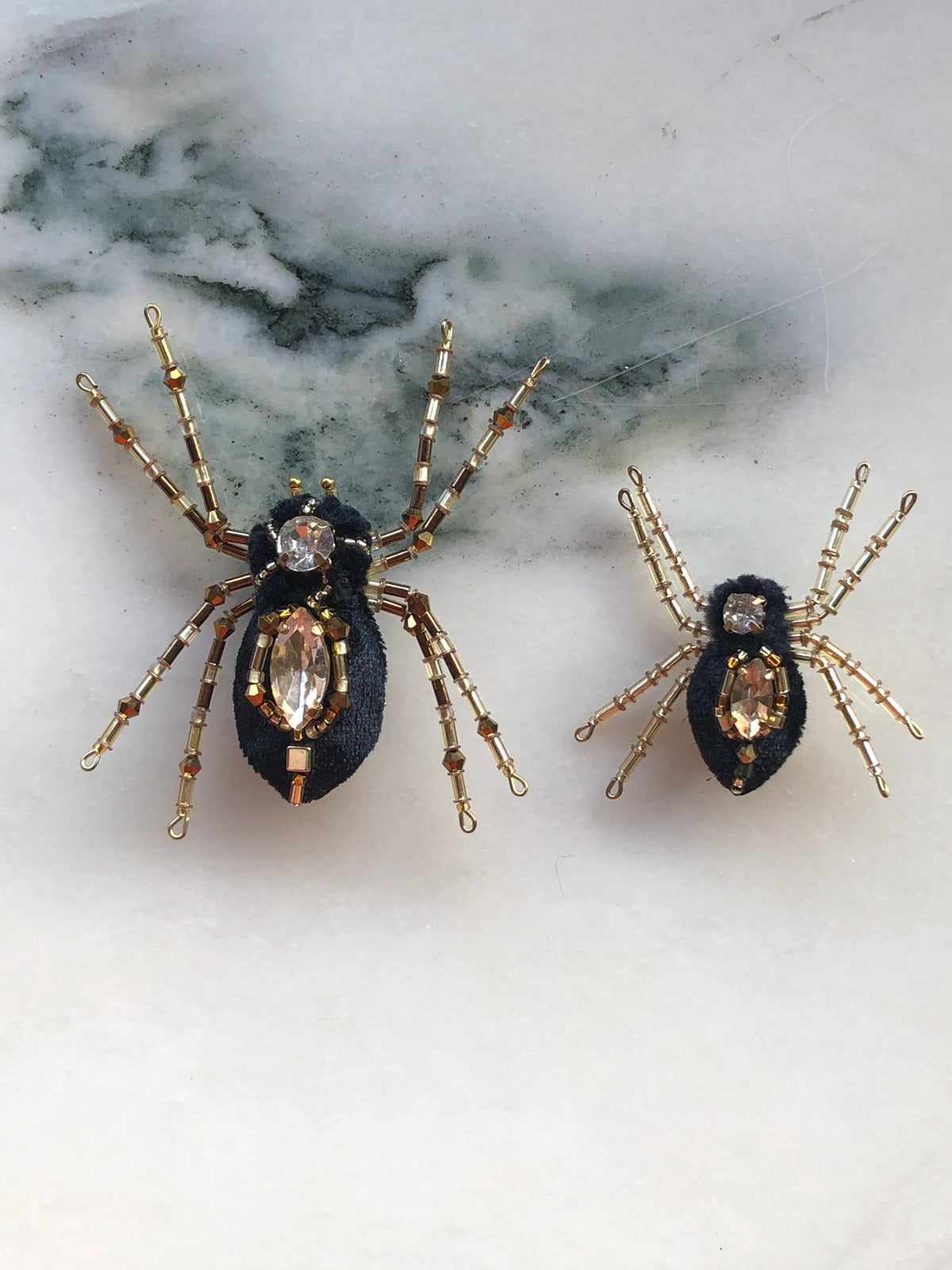 Broochella Ragno e Ragnetto Spider Brooch Pin Set featuring two black textile spiders embellished with gold-tone beadwork and faceted crystal details. Pictured on a white marble surface