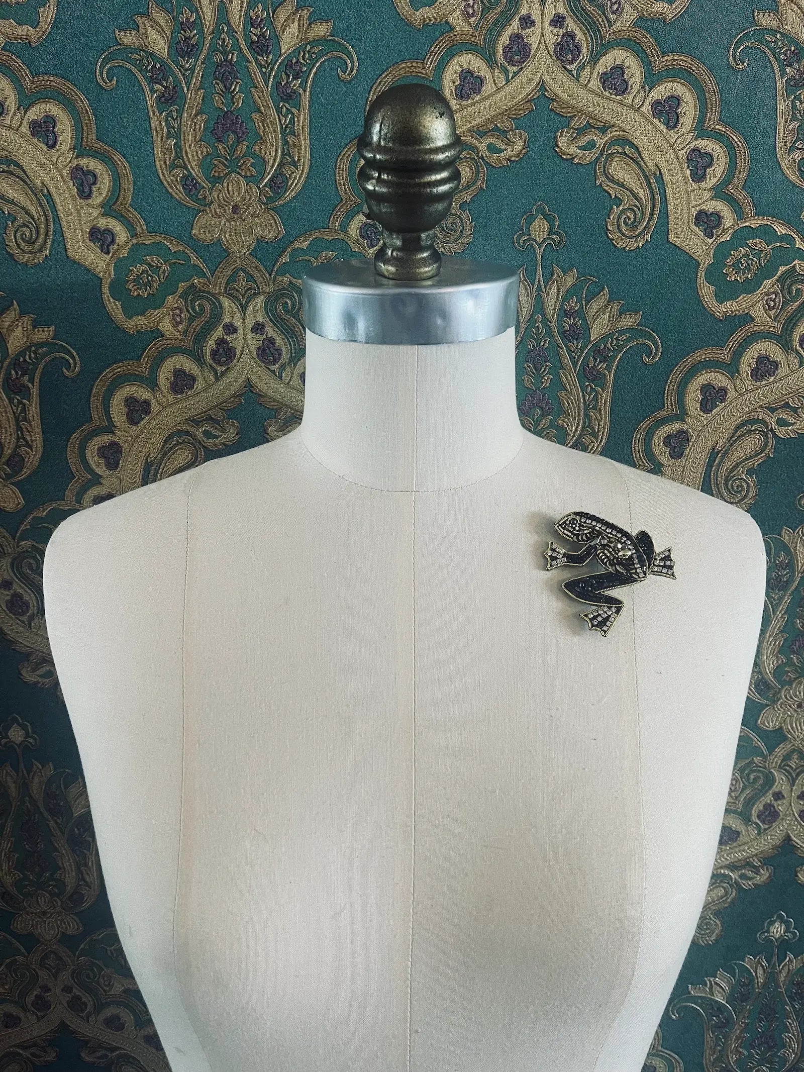 broochella hand beaded black and gold frog brooch. Pictured on a mannequin. The background is green and gold damask wallpaper