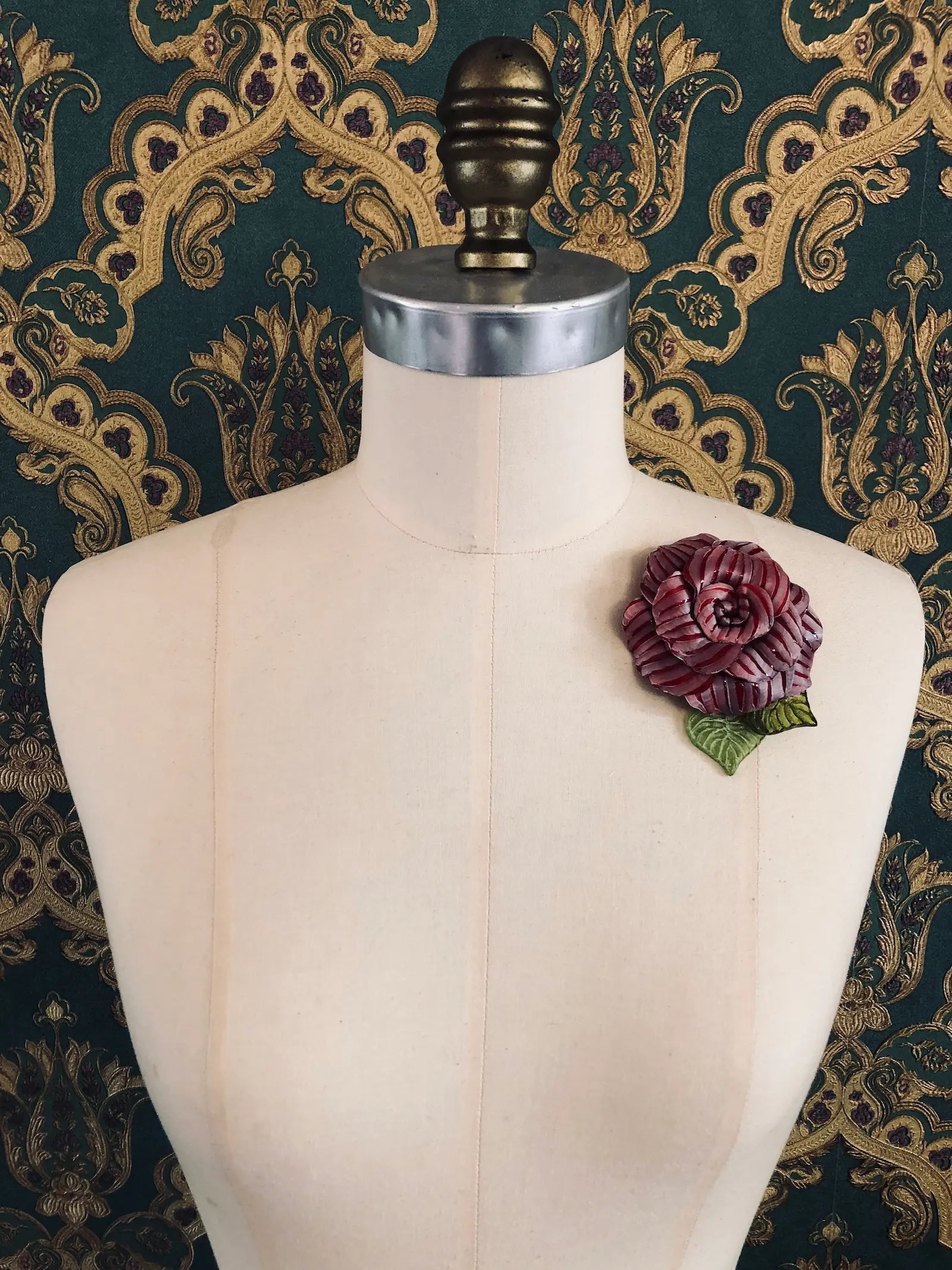 A Broochella x Fabrice Paris red moulded plastic rose brooch with green leaves, pinned to the chest of a cream-coloured dress form against a dark green and gold patterned damask backdrop.