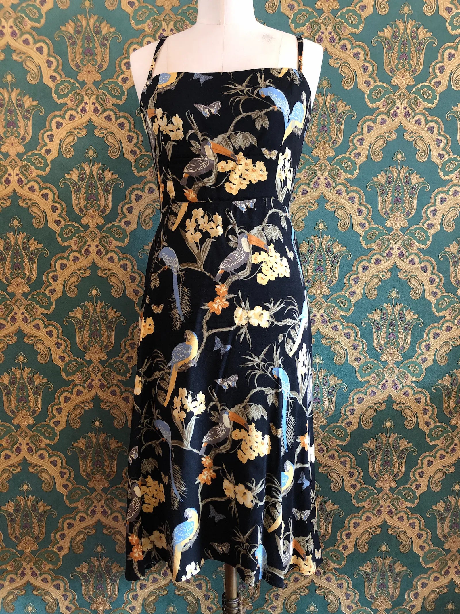 Broochella Reformation black midi dress with colourful tropical birds, butterflies and floral print, size 10. Pictured on a mannequin. The background is green and gold damask wallpaper