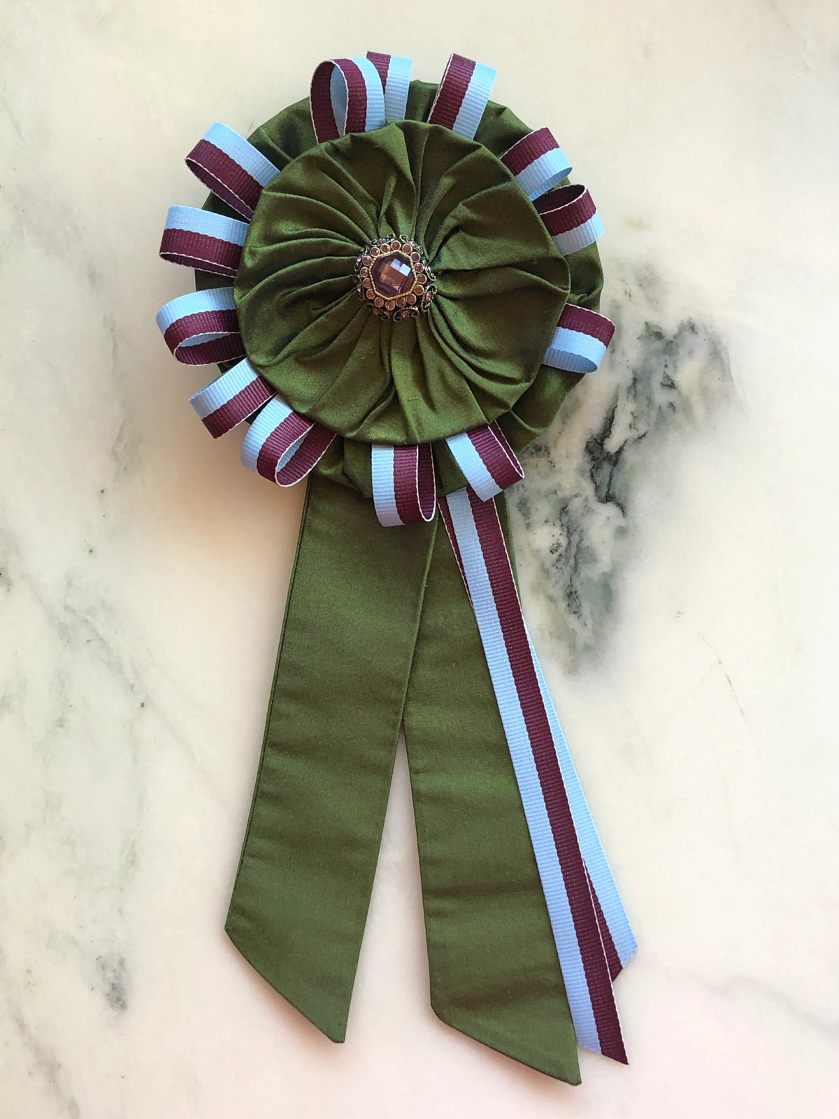 Broochella Regalia Ribbon Brooch in moss green, a handmade designer statement brooch featuring a sculptural fabric rosette, burgundy and pale blue ribbon loops, long ribbon tails and a jewelled centre embellishment, photographed on white marble.