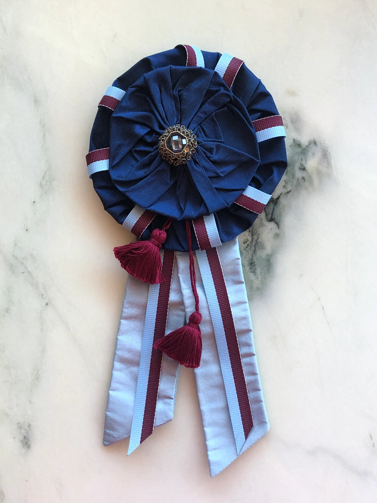 Broochella Regalia Ribbon Brooch in navy blue, a handmade designer statement brooch featuring a layered fabric rosette, burgundy and pale blue striped ribbon, long satin tails, burgundy tassels and an ornate jewelled centre, photographed on white marble.