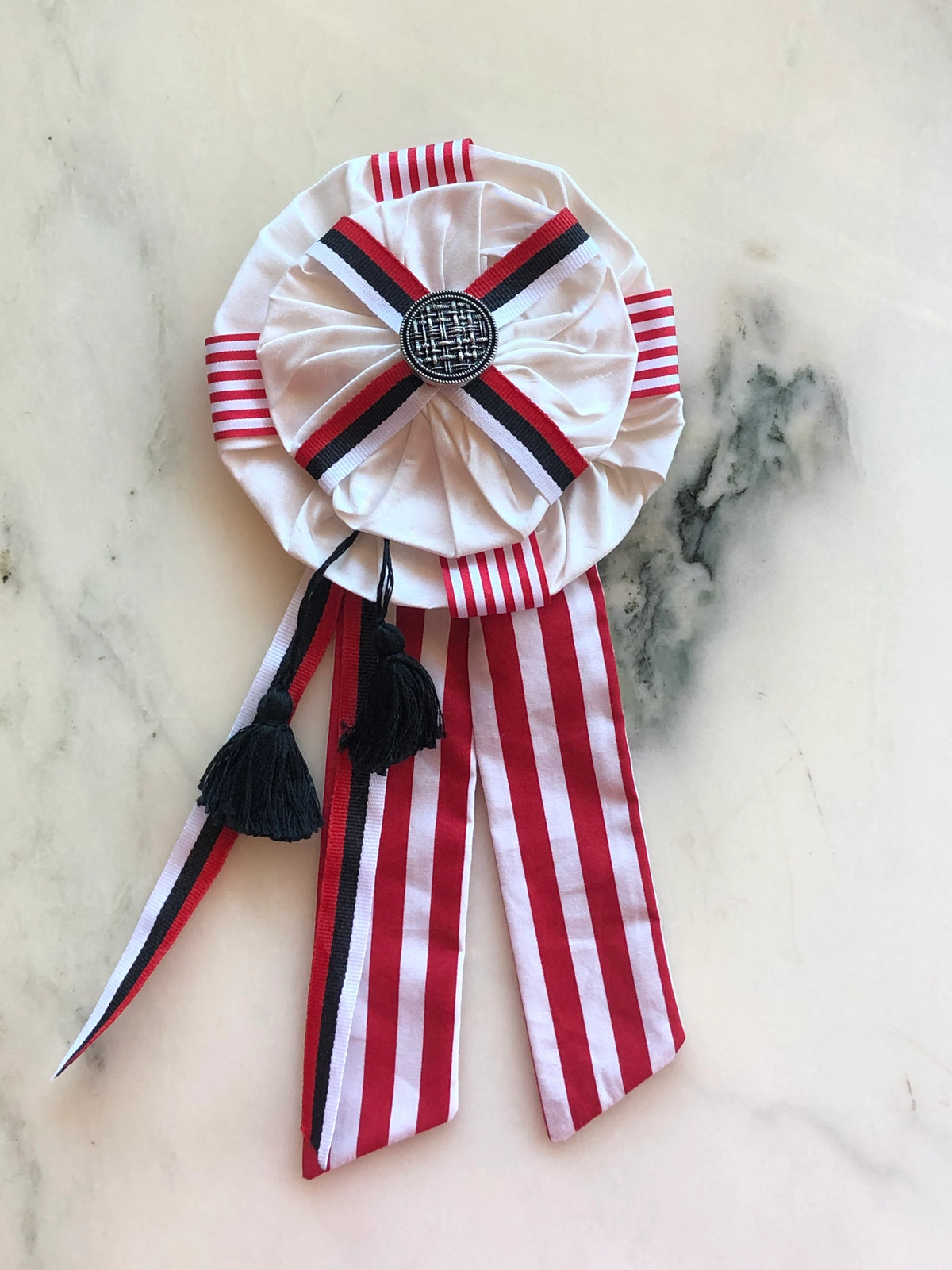 Broochella Regalia Ribbon Brooch in red and white, a handmade designer statement brooch featuring layered ivory fabric rosettes, bold striped ribbon, black detailing, tassels and an ornate centre embellishment, photographed on white marble.
