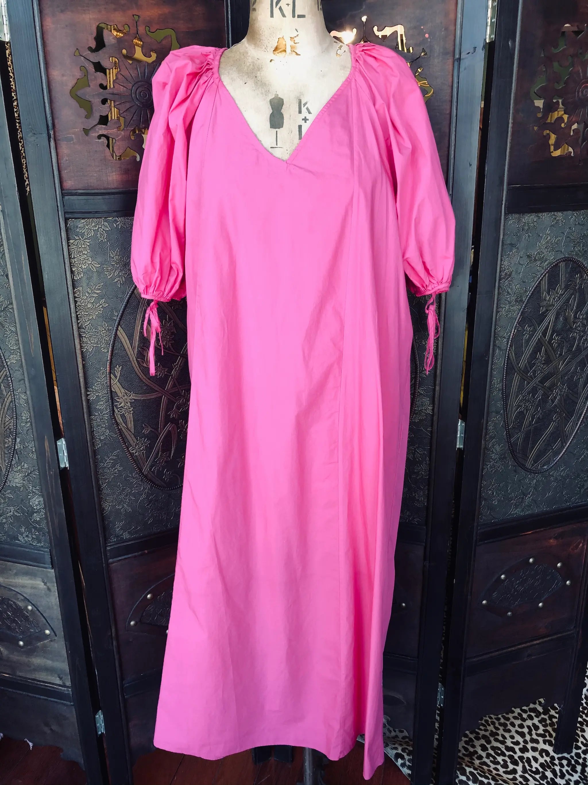 Broochella, Rhode, XS midi dress. It is bright pink, 100% Cotton, with a V-neckline, Short puff sleeves with tie cuffs and a relaxed fit. It is pictured on a vintage mannequin. The background is an ornate, vintage, dark brown wood screen. 