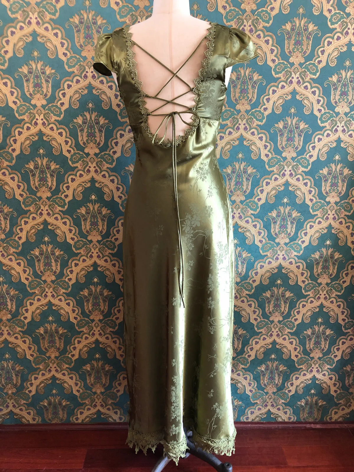 Broochella Rihoas olive-green floral jacquard satin maxi dress with lace trim and an open tie back, size Medium. Pictured on a mannequin. The background is green and gold damask wallpaper