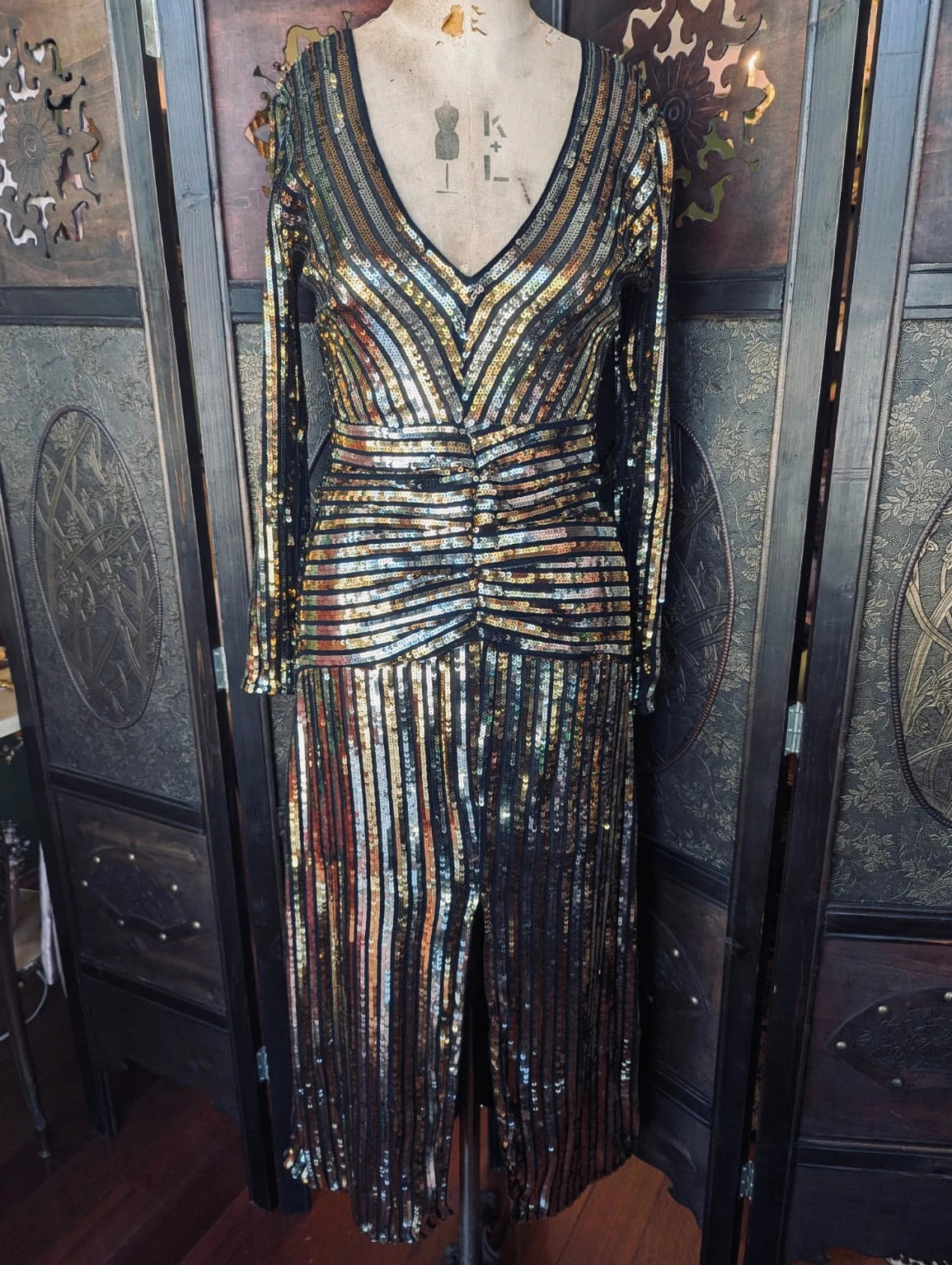 Broochella Rixo Black, long sleeve midi dress with silver and gold sequin striped detail. It is presented on a vintage mannequin. The background is an ornate, dark wood room divider. 