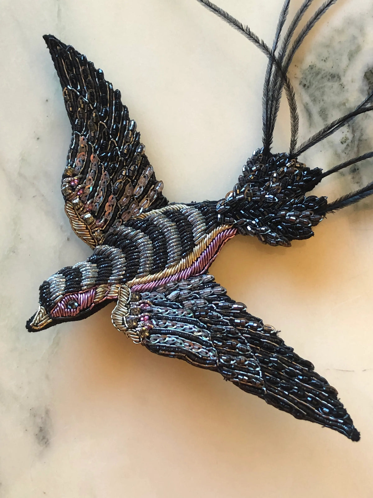 Broochella Rondine Bird Brooch Pin featuring a dramatic flying bird hand embellished with black, bronze and metallic beads and sequins, finished with long sweeping black feather tail details on a cream marble background.
