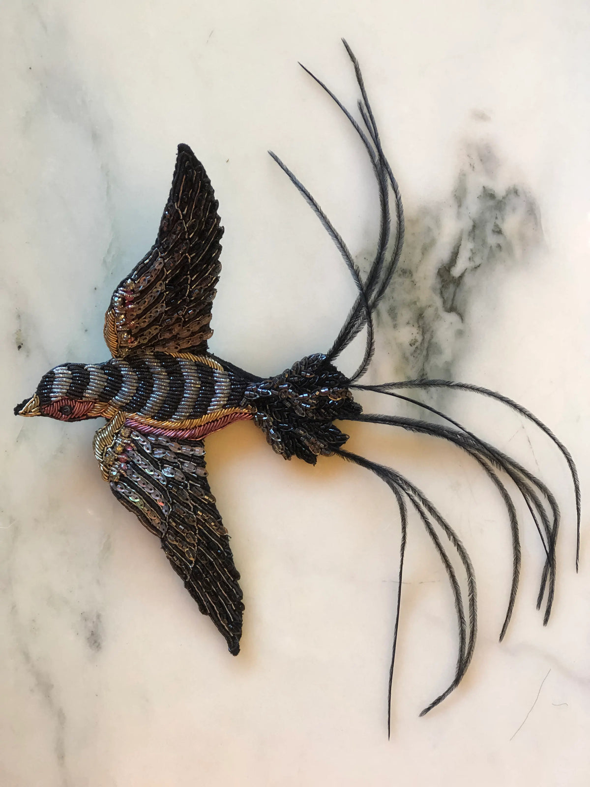 Broochella Rondine Bird Brooch Pin featuring a dramatic flying bird hand embellished with black, bronze and metallic beads and sequins, finished with long sweeping black feather tail details on a cream marble background.