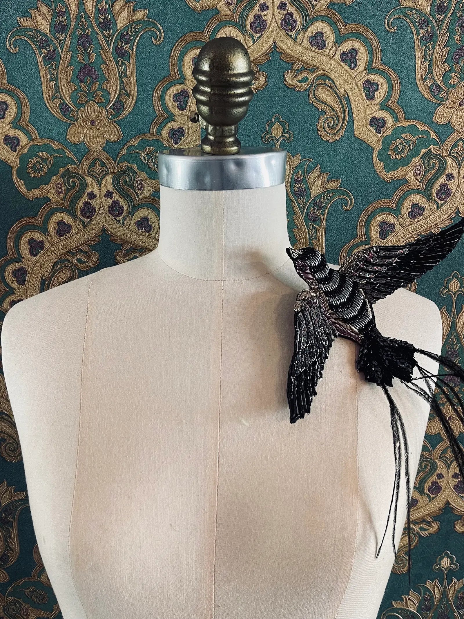 Broochella Rondine handmade hand beaded pink and black figural bird brooch. Styled on a mannequin. The background is green and gold damask wallpaper
