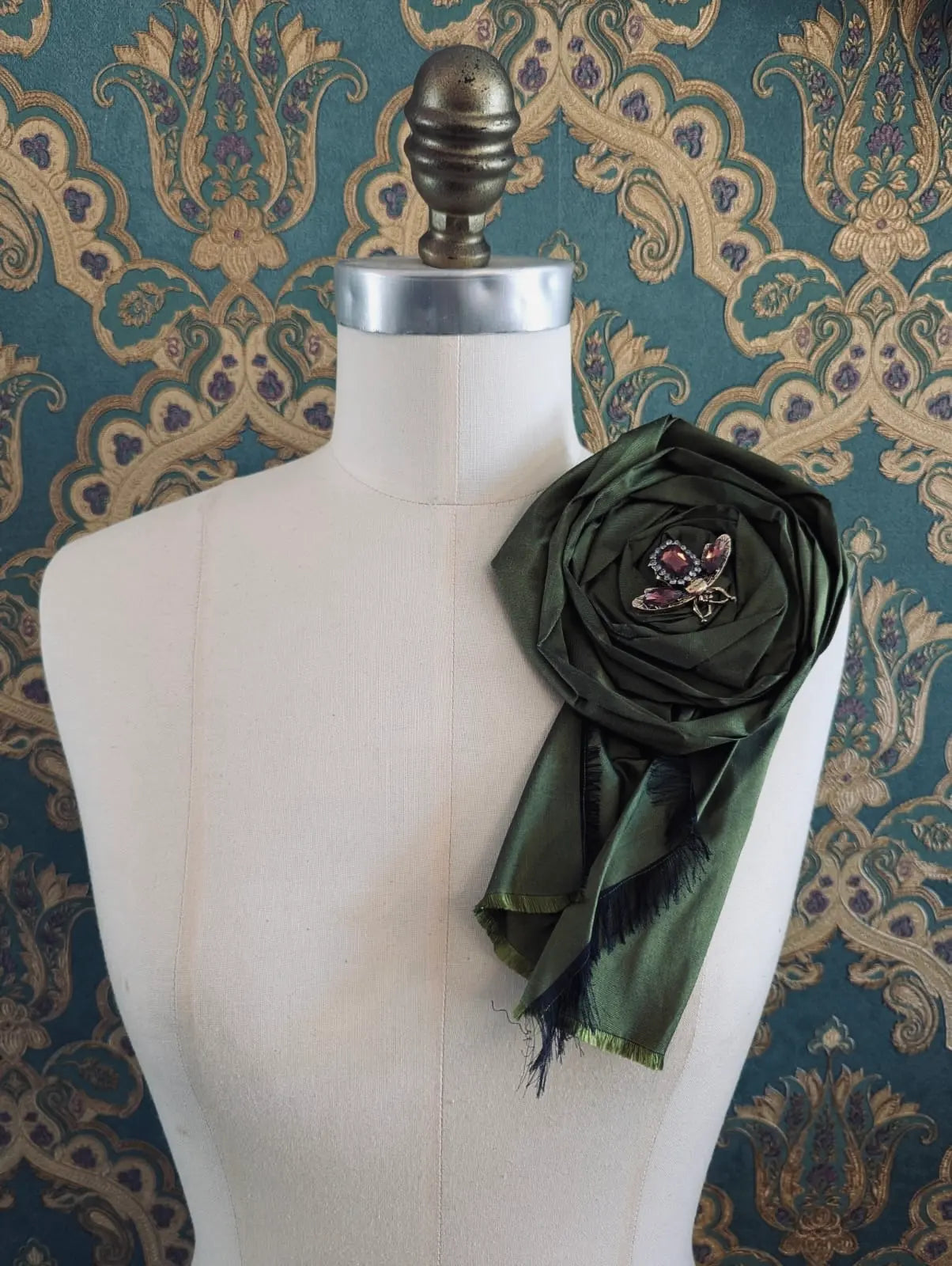 Broochella rosa ingioiellata green handmade designer silk silk flower brooch with rhinestone insect embellishment. styled on a mannequin. The background is green and gold damask wallpaper