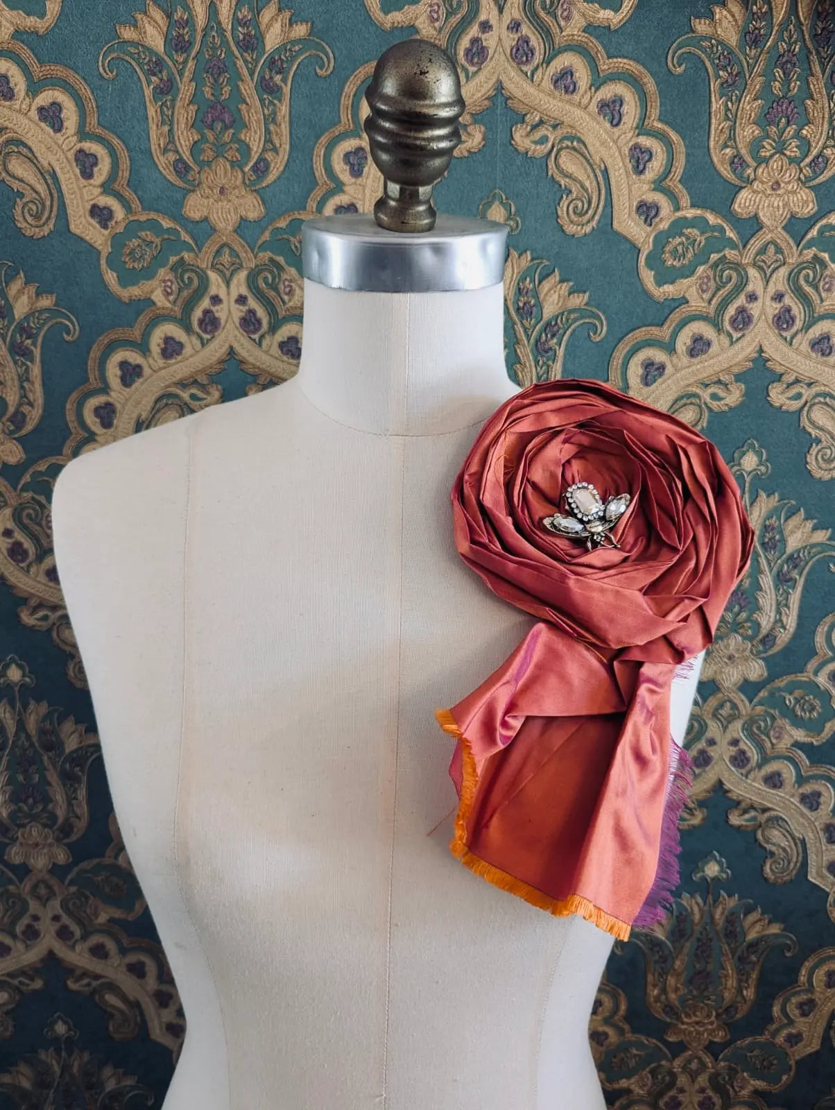 Broochella rosa ingioiellata orange handmade designer silk silk flower brooch with rhinestone insect embellishment. styled on a mannequin. The background is green and gold damask wallpaper