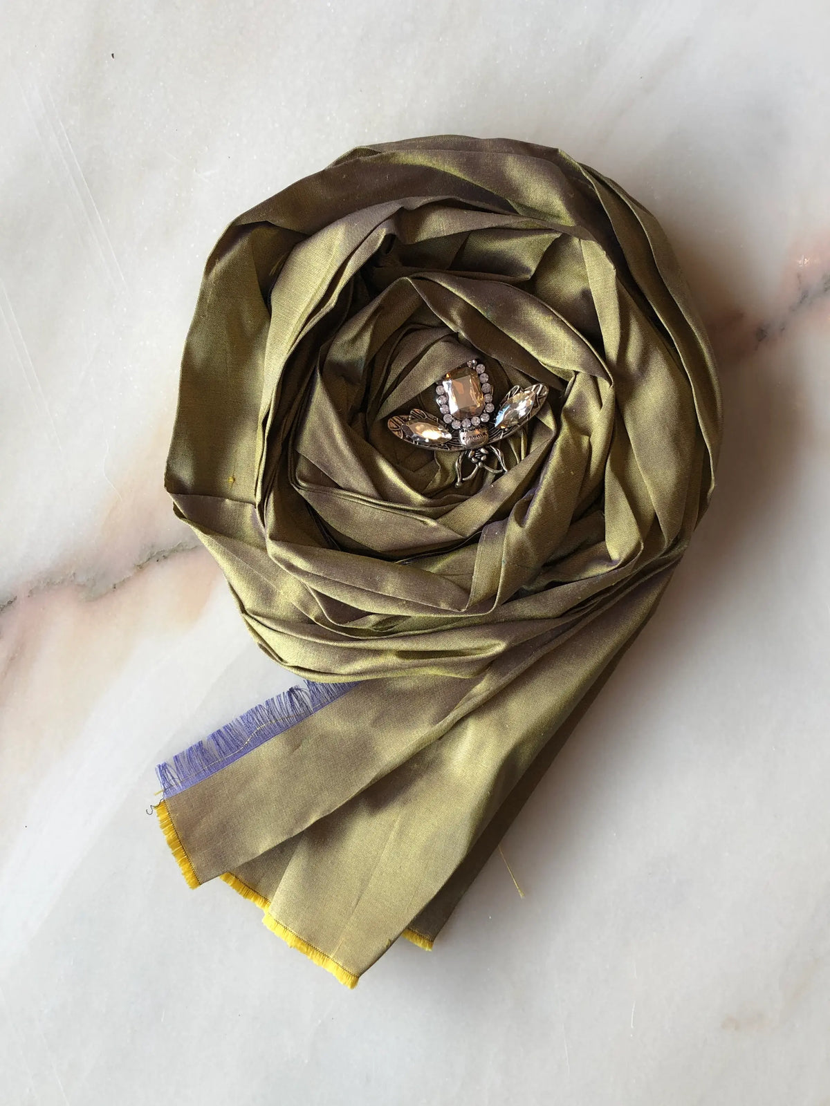 Broochella Rosa Ingioiellata Large handmade designer flower brooch in shimmering antique gold silk, sculpted into an oversized rose and finished with a crystal jewelled insect centrepiece, photographed on cream marble with natural grey veining.
