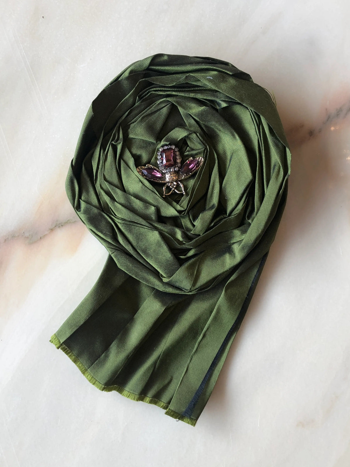 Broochella Rosa Ingioiellata Large handmade designer flower brooch in deep green silk, sculpted into an oversized rose and finished with a purple crystal jewelled insect centrepiece, photographed on cream marble with natural grey veining.