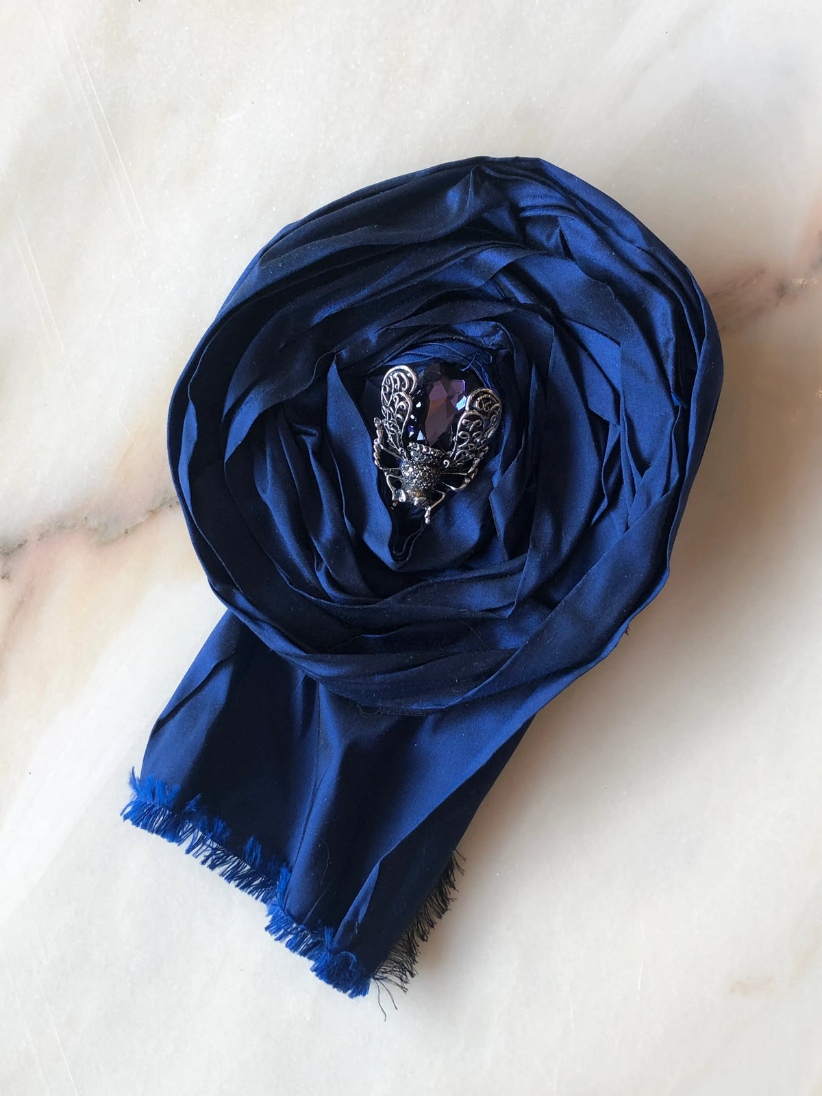 Broochella Rosa Ingioiellata Large handmade designer flower brooch in deep navy silk, sculpted into an oversized rose and finished with a dark crystal jewelled insect centrepiece, photographed on cream marble with natural grey veining.