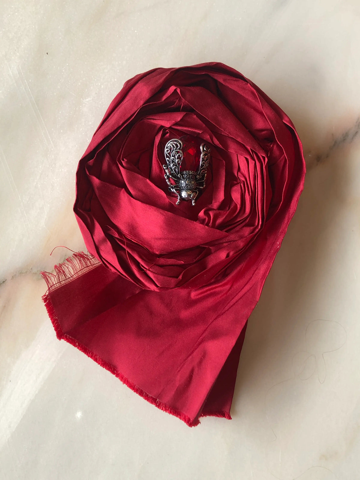 Broochella Rosa Ingioiellata Large handmade designer flower brooch in rich red silk, sculpted into an oversized rose and finished with a jewelled insect centrepiece, photographed on cream marble with natural grey veining.
