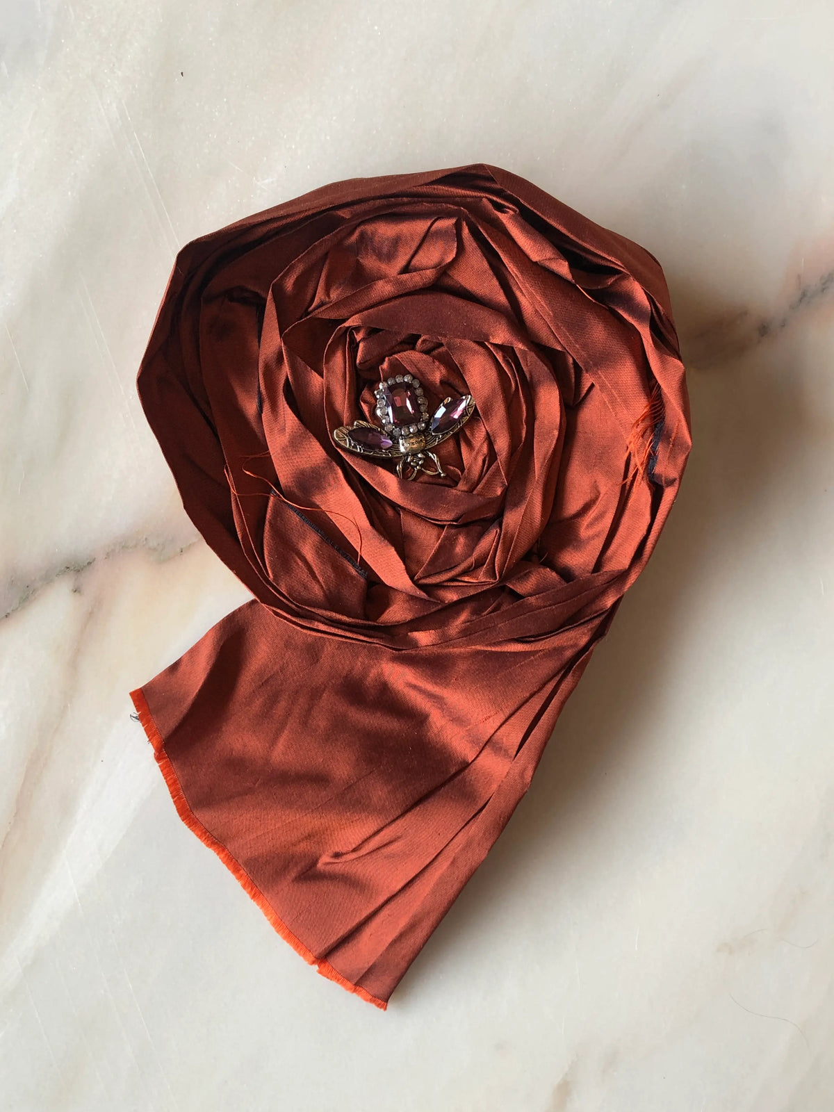 Broochella Rosa Ingioiellata Large handmade designer flower brooch in lustrous rust silk, sculpted into an oversized rose and finished with a crystal jewelled insect centrepiece, photographed on cream marble with natural grey veining.