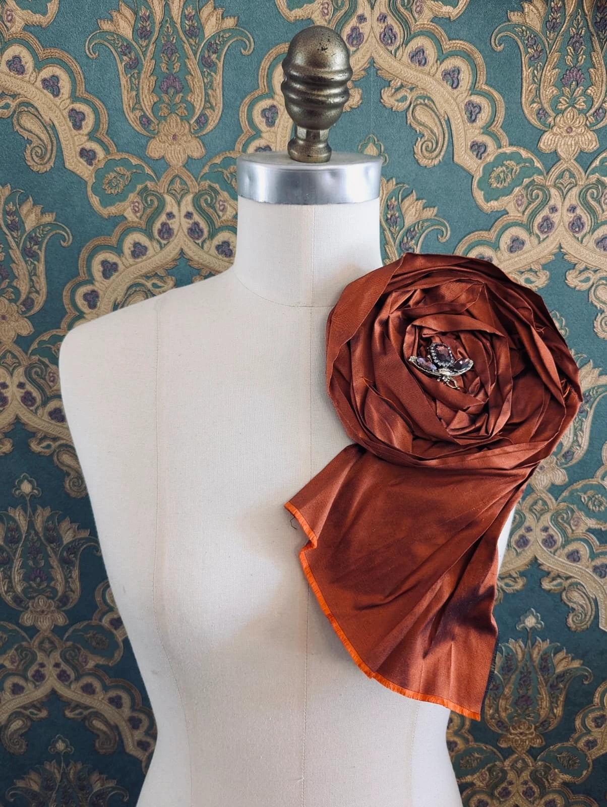 Broochella rosa ingioiellata large rust handmade silk flower brooch with an insect brooch embellishment in the centre. It is pinned to a mannequin. The background is green and gold damask wallpaper.