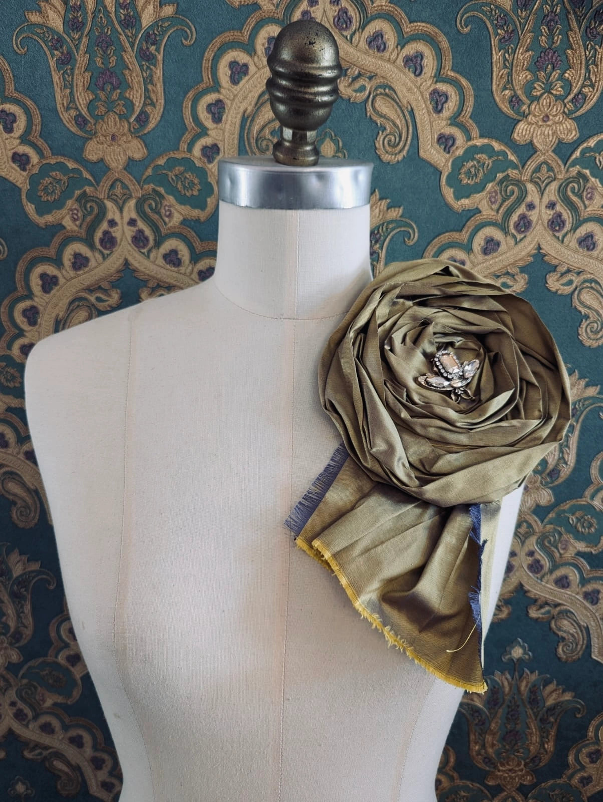 Broochella rosa ingioiellata large gold handmade silk flower brooch with an insect brooch embellishment in the centre. It is pinned to a mannequin. The background is green and gold damask wallpaper.