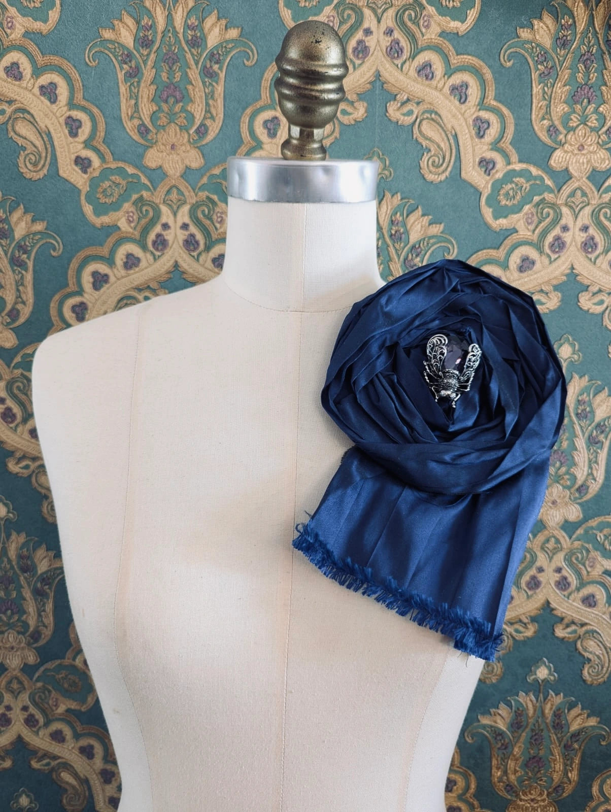 Broochella rosa ingioiellata large navy handmade silk flower brooch with an insect brooch embellishment in the centre. It is pinned to a mannequin. The background is green and gold damask wallpaper.