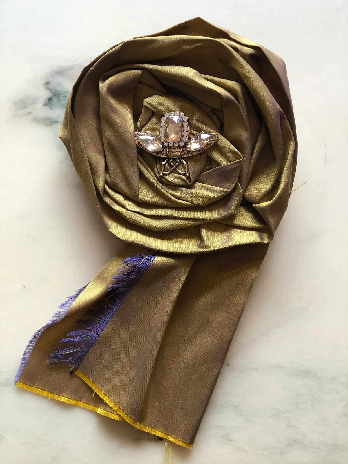 Broochella Rosa Ingioiellata Flower Brooch Pin - Small in rich gold silk, styled with a vintage blue crystal and silver-tone brooch at its centre on a cream marble background.