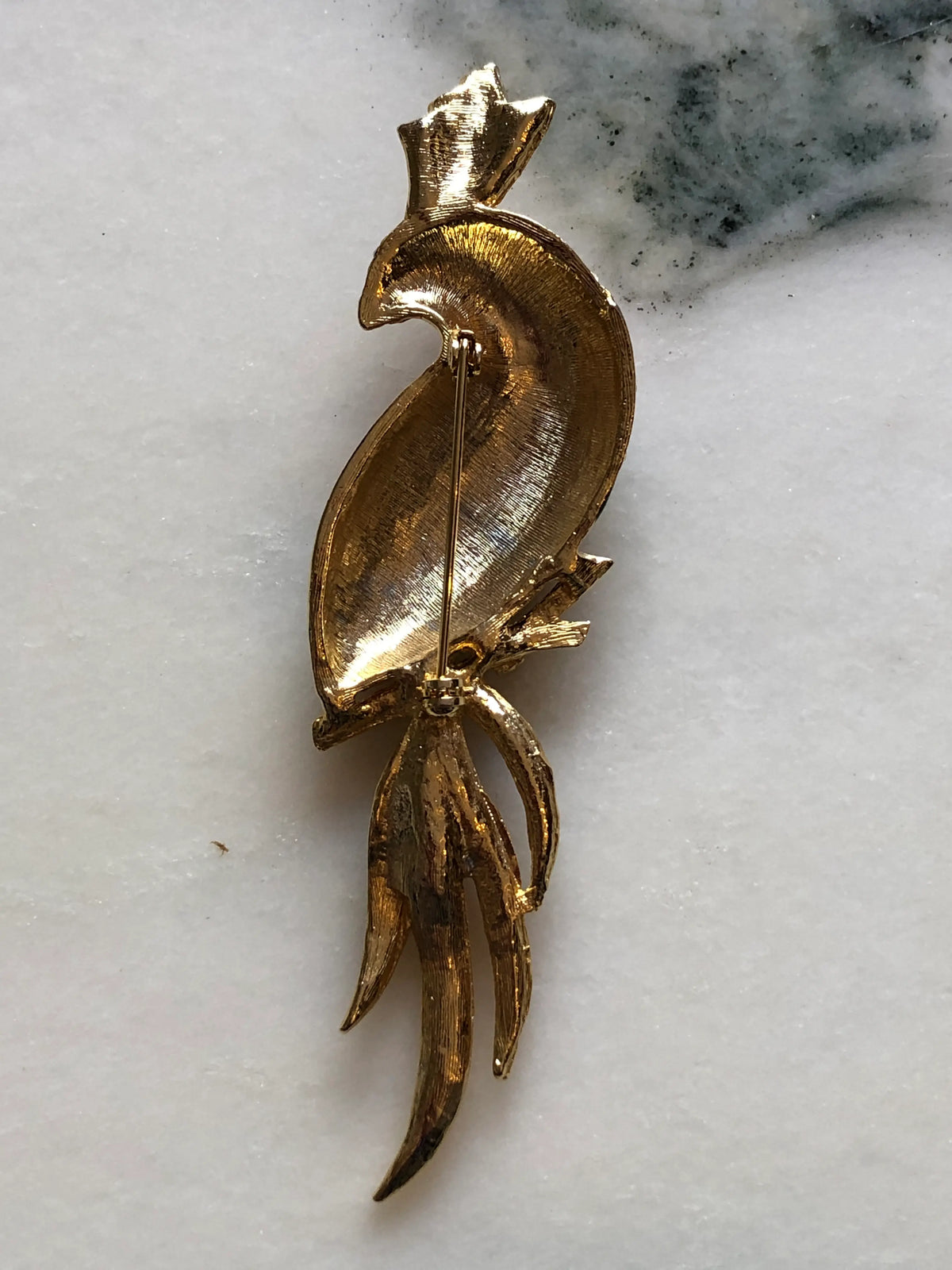 Broochella Rosalia Vintage Enamel Parrot Brooch featuring red, green and navy enamel, gold-tone detailing and clear crystals with a long multicoloured tail on a cream and grey marble background.