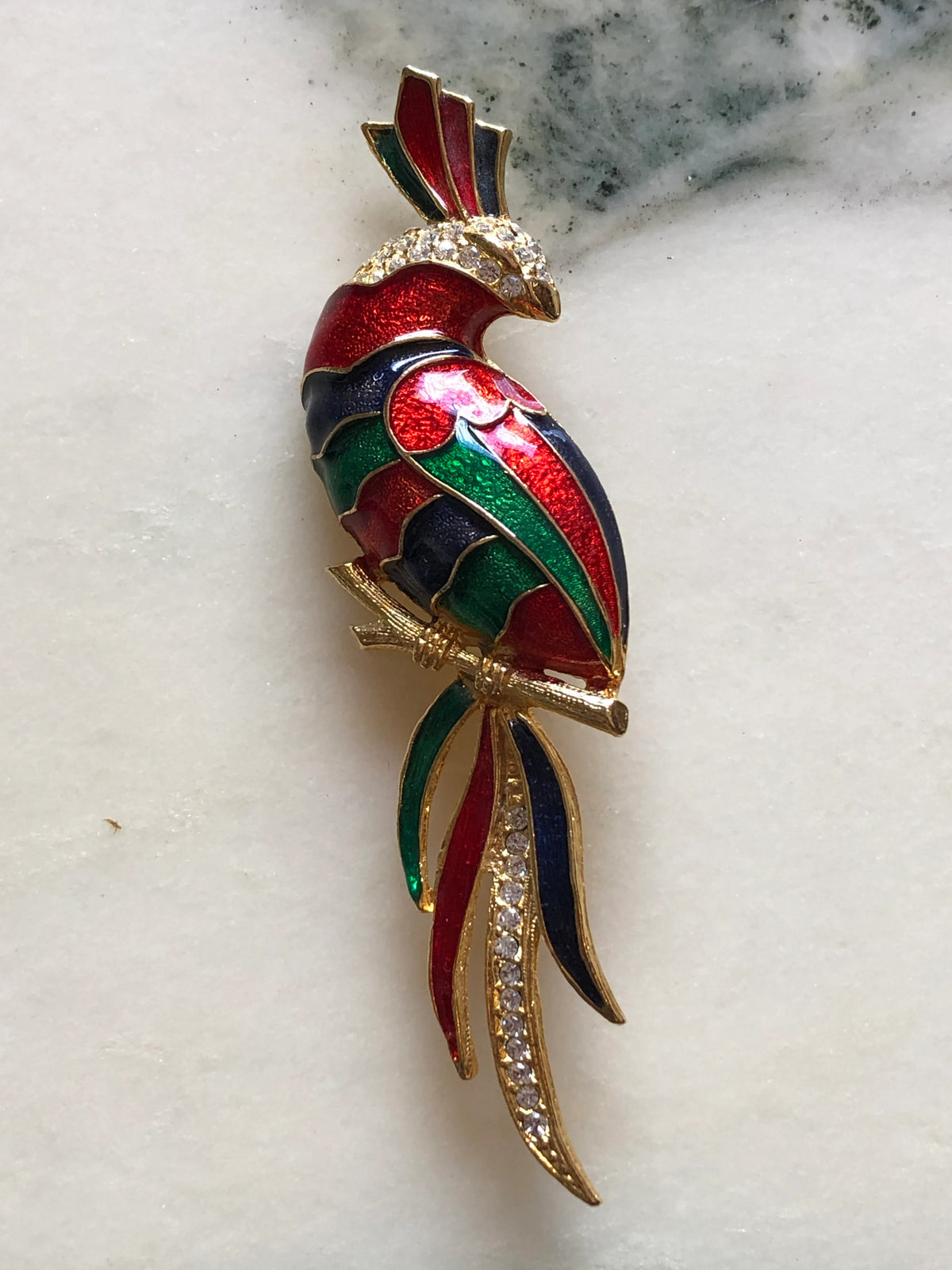 Broochella Rosalia Vintage Enamel Parrot Brooch featuring red, green and navy enamel, gold-tone detailing and clear crystals with a long multicoloured tail on a cream and grey marble background.