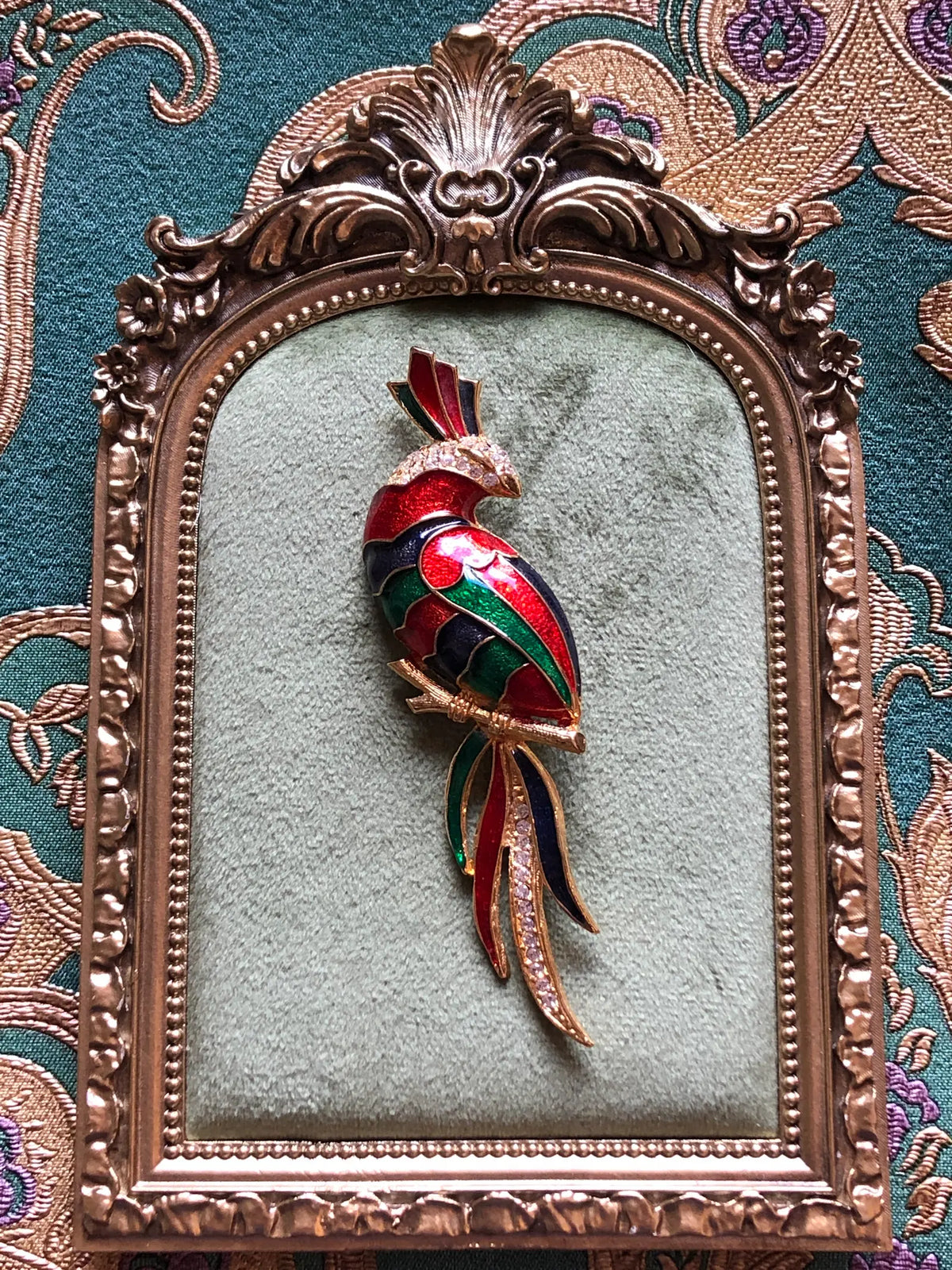 Broochella Rosalia Vintage Enamel Parrot Brooch featuring red, green and navy enamel, gold-tone detailing and clear crystals with a long multicoloured tail. Pictured on a green velvet tray with an ornate gold frame. The background is green and gold damask wallpaper
