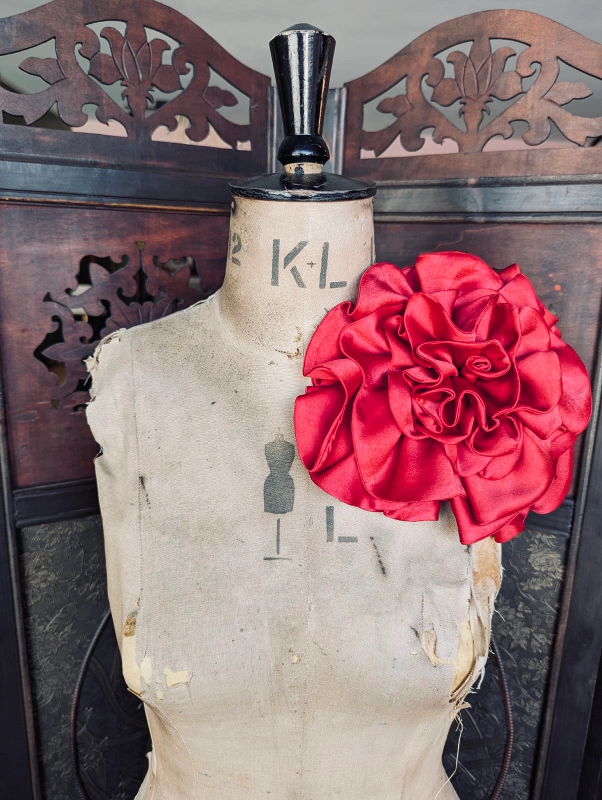 Broochella rose de minuit large, red, handmade, satin flower brooch. It is pinned to a vintage mannequin. In the background is a decorative wooden screen.