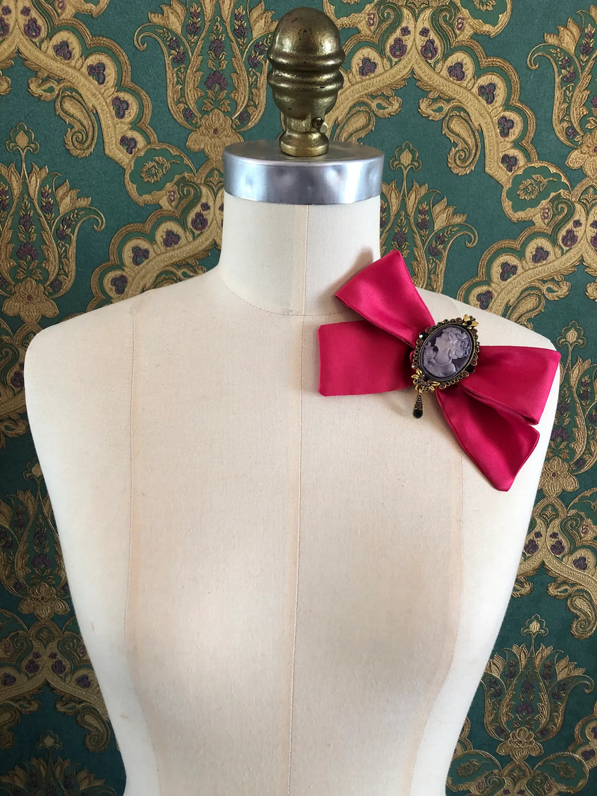 Broochella Rosée Camée Pink Cameo Bow Brooch featuring a vivid pink satin bow and lilac cameo centre, styled on a cream mannequin against ornate green, gold and purple patterned wallpaper.