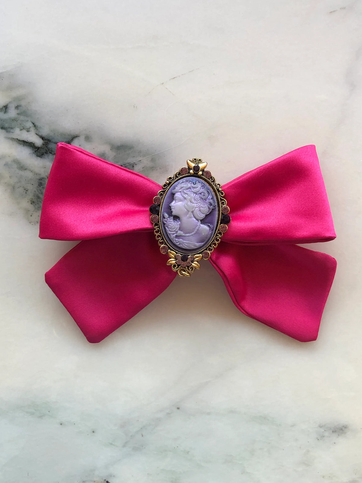 Broochella Rosée Camée Pink Cameo Bow Brooch featuring a vivid pink satin bow with an ornate gold-tone setting and lilac portrait cameo, photographed on white marble with soft grey-green veining.