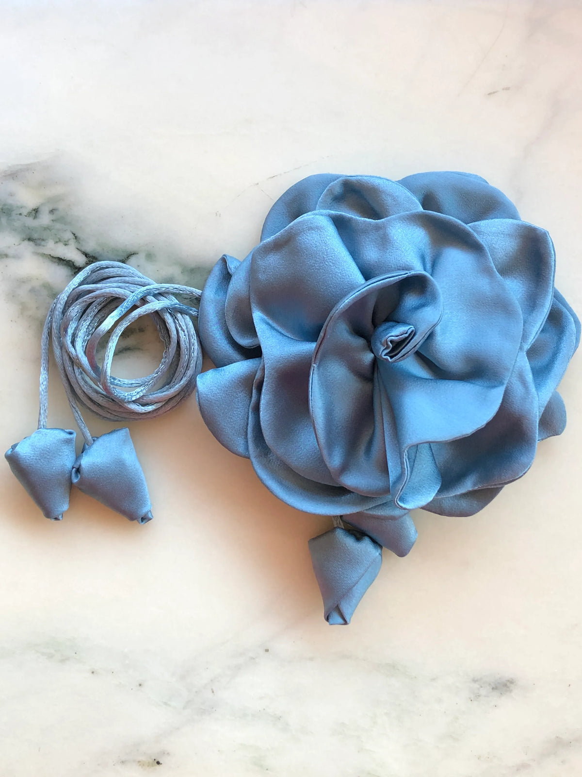 Broochella Rosetta blue flower choker and brooch with sculptural satin petals, matching tie cords and fabric buds, photographed on white marble with grey veining.