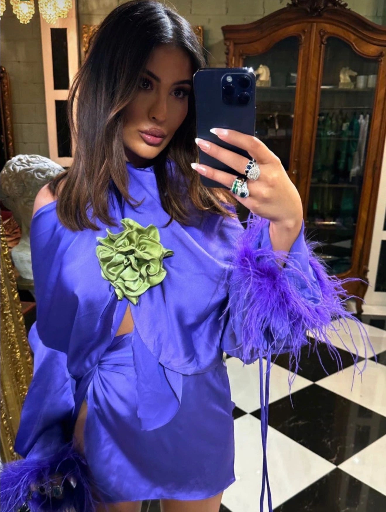 A woman takes a mirror selfie wearing a vibrant purple off-the-shoulder dress with a large green broochella rosetta fabric flower brooch on the chest and purple feathered cuffs. She is wearing multiple rings on her hand, which holds a black smartphone.