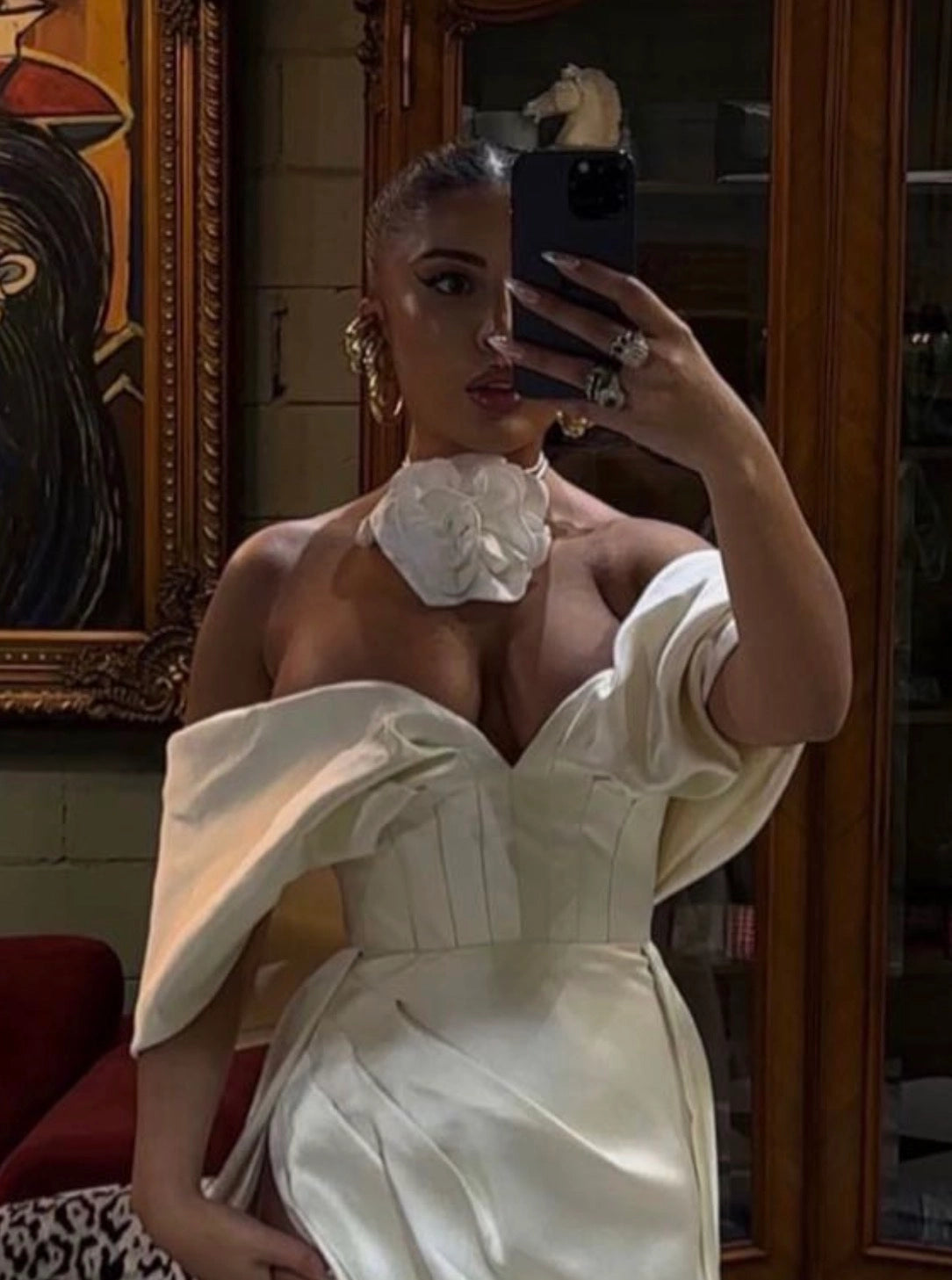 A woman takes a mirror selfie wearing a structured off-the-shoulder ivory dress with a Broochella rosetta flower choker in ivory, set against an indoor background with wooden furniture and artwork.