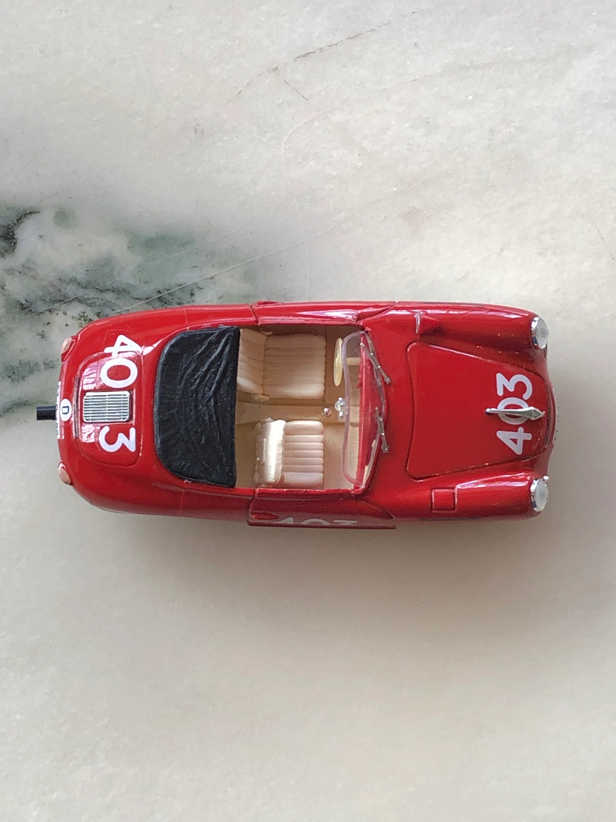 Broochella Rosso Mille No.403 Vintage Convertible Racing Car Brooch photographed from above, showing its glossy red body, white number 403 decals, open cream interior, black folded roof and detailed windscreen.
