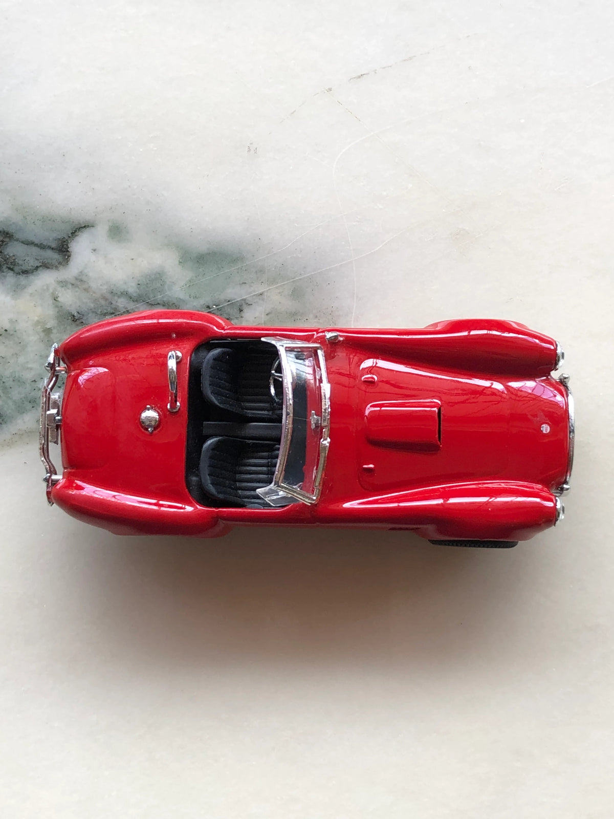 Broochella Rosso Roadster No.427 Vintage Convertible Racing Car Brooch photographed from above, showcasing its bright red body, open cockpit with two black seats, chrome-framed windscreen, sculpted bonnet and silver detailing