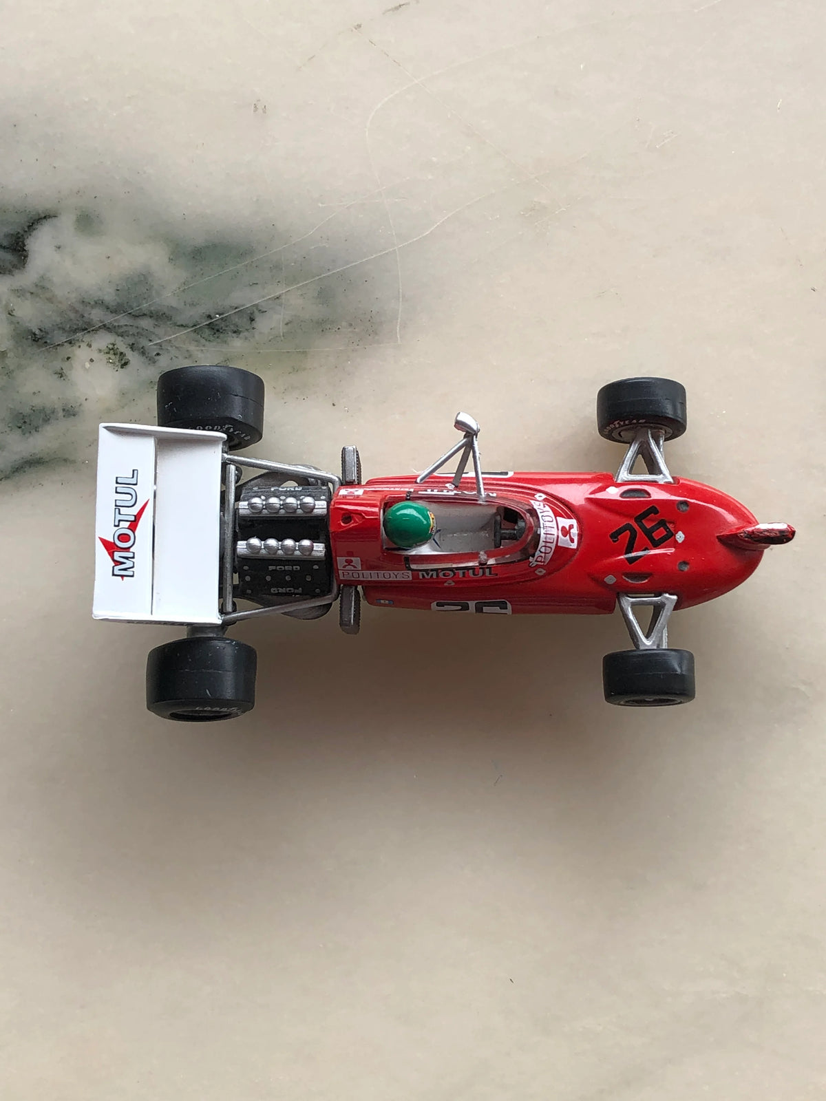 Broochella Rosso Turbo No.26 Vintage Formula Racing Car Brooch viewed from above, featuring a bright red body, black number 26 decals, green racing helmet, exposed Ford engine, black tyres and white Motul rear wing.