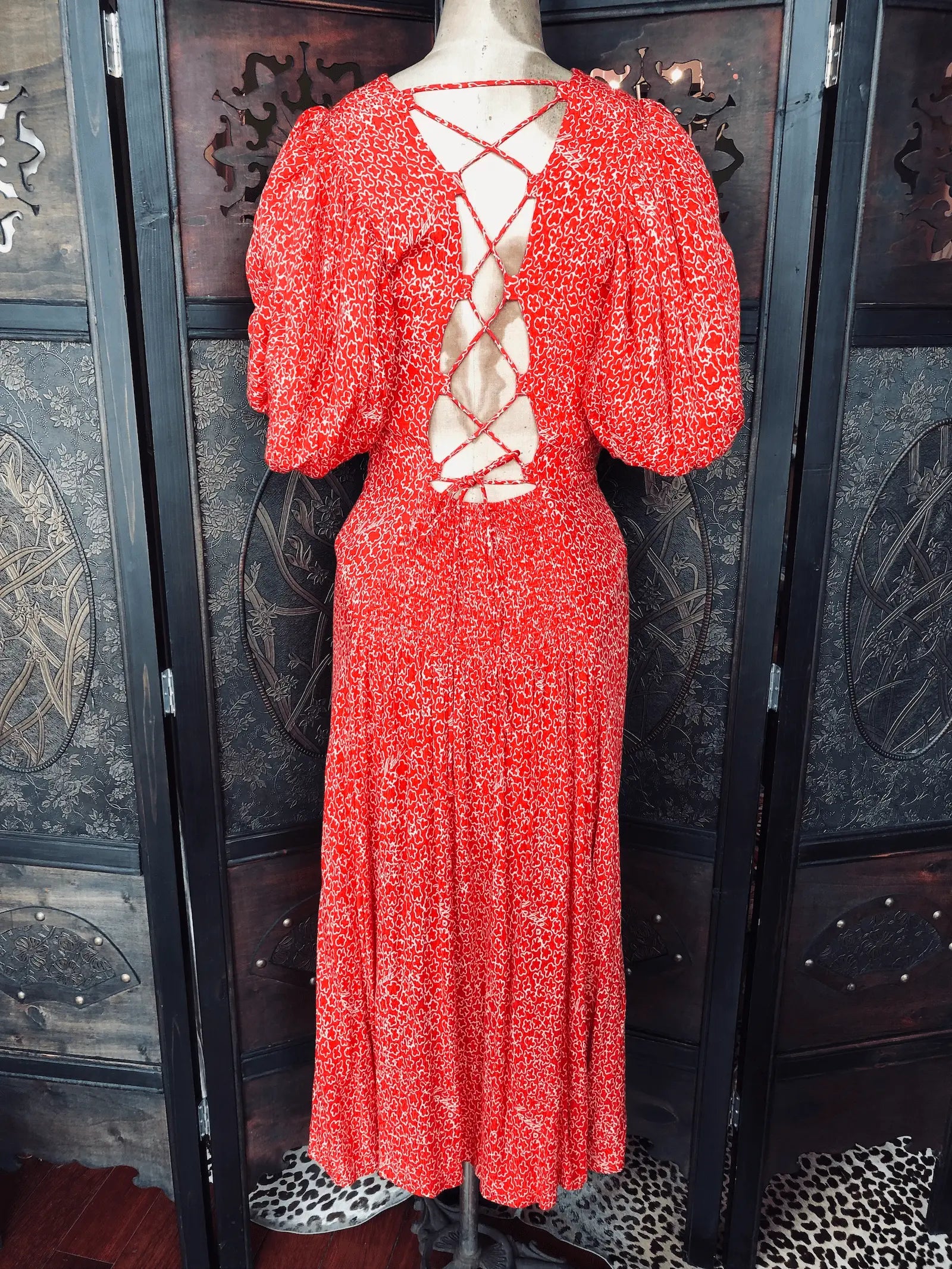 A Broochella Rotate red and white patterned midi dress with puff sleeves and a criss-cross lace-up open back, displayed on a mannequin in front of a dark wooden folding screen.