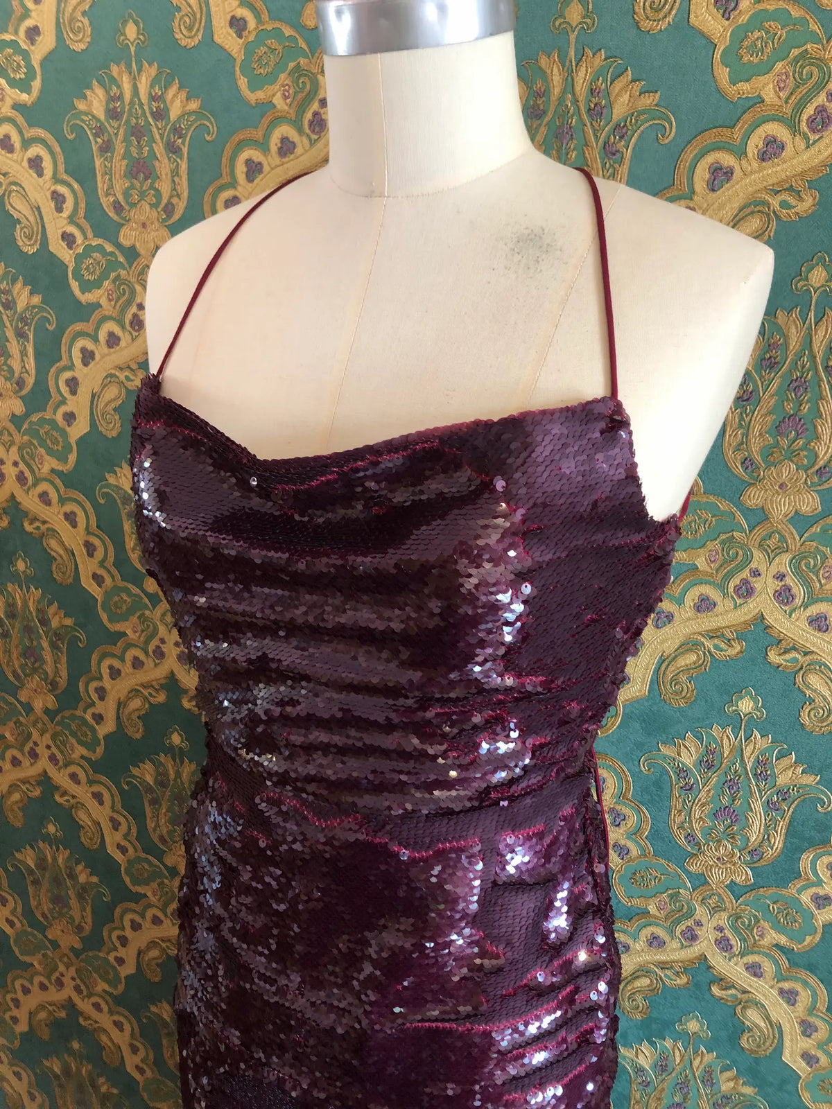 The side view of Broochella Sabina Musáyev Zarya midi dress in Ruby Wine with tonal sequins, cowl neckline and lace-up back, size XS. Pictures on a mannequin. The background is green and gold damask wallpaper