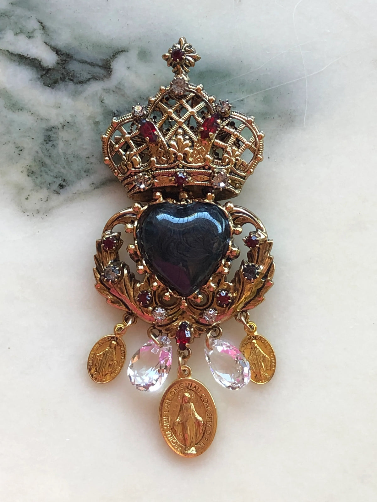 Broochella Sacro Cuore Agatha Brooch in ornate gold with a black heart, jewelled crown, crystal drops and devotional medallions, photographed on a white marble background.