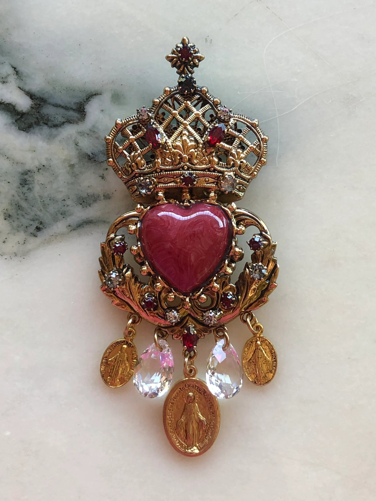 Broochella Sacro Cuore Agatha Brooch in ornate gold with a red heart, jewelled crown, crystal drops and devotional medallions, photographed on a white marble background.