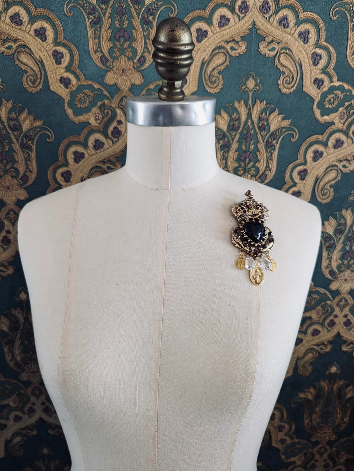 Broochella Sacro cuore agatha gold and black designer crown and heart shaped brooch. Pinned to a mannequin. The background is green and gold damask wallpaper