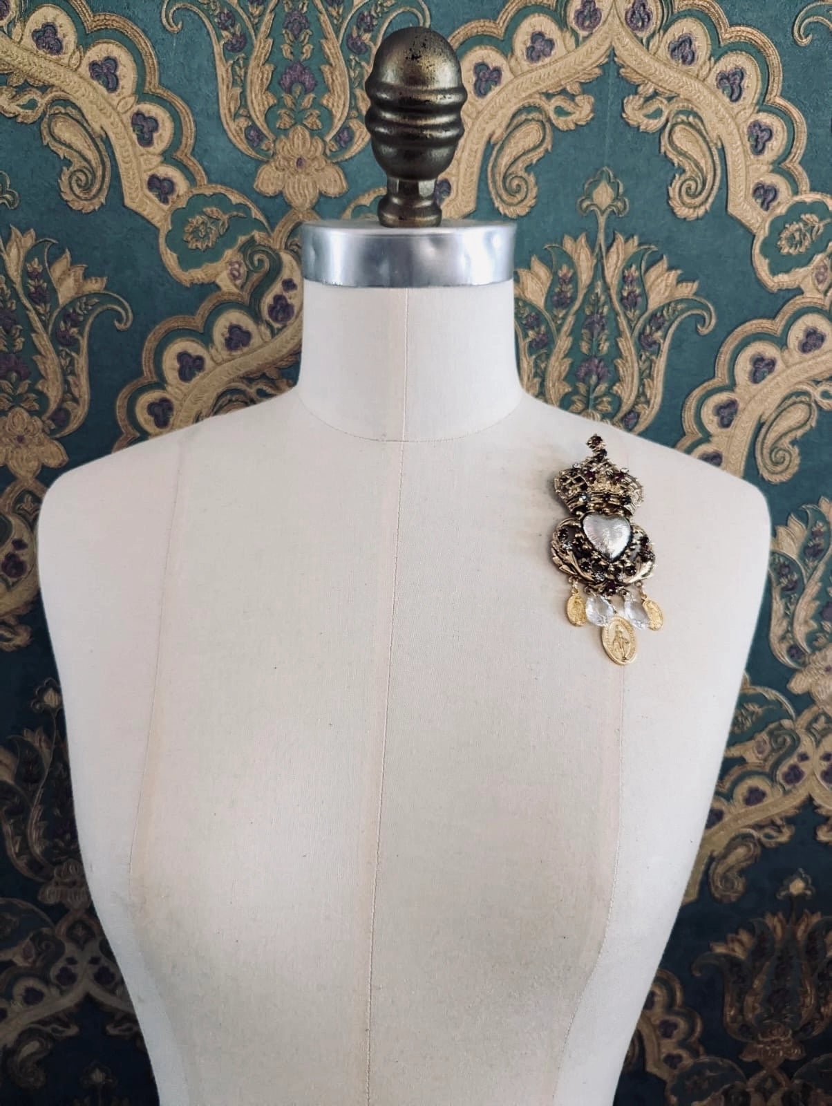 Broochella Sacro cuore agatha gold designer crown and heart shaped brooch. Pinned to a mannequin. The background is green and gold damask wallpaper