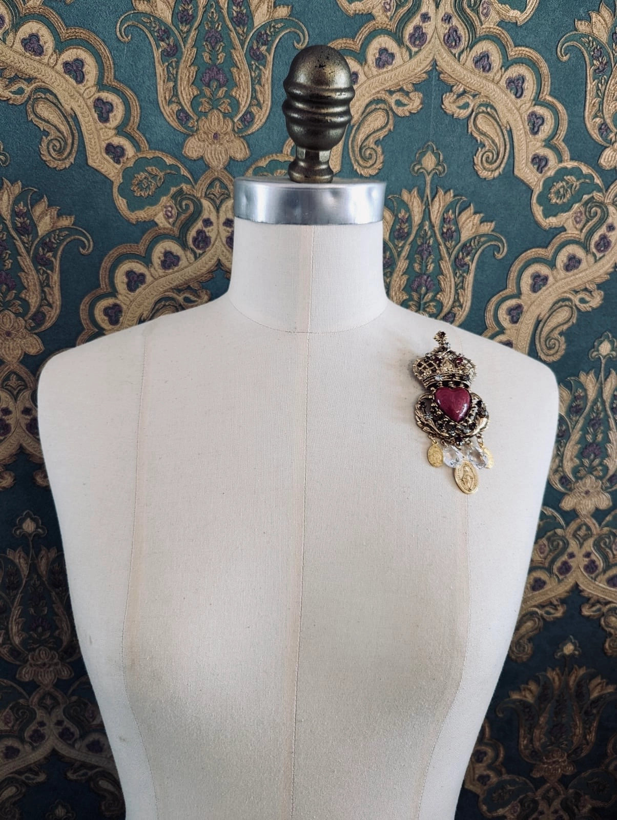 Broochella Sacro cuore agatha gold and red designer crown and heart shaped brooch. Pinned to a mannequin. The background is green and gold damask wallpaper